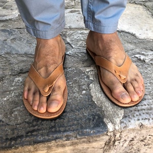 Pode incluir: Um par de chinelos de couro castanho claro. As sand&aacute;lias t&ecirc;m um design de tanga com uma tira entre os dedos. As solas s&atilde;o castanhas mais escuras. As sand&aacute;lias s&atilde;o usadas com cal&ccedil;as cinza claro.