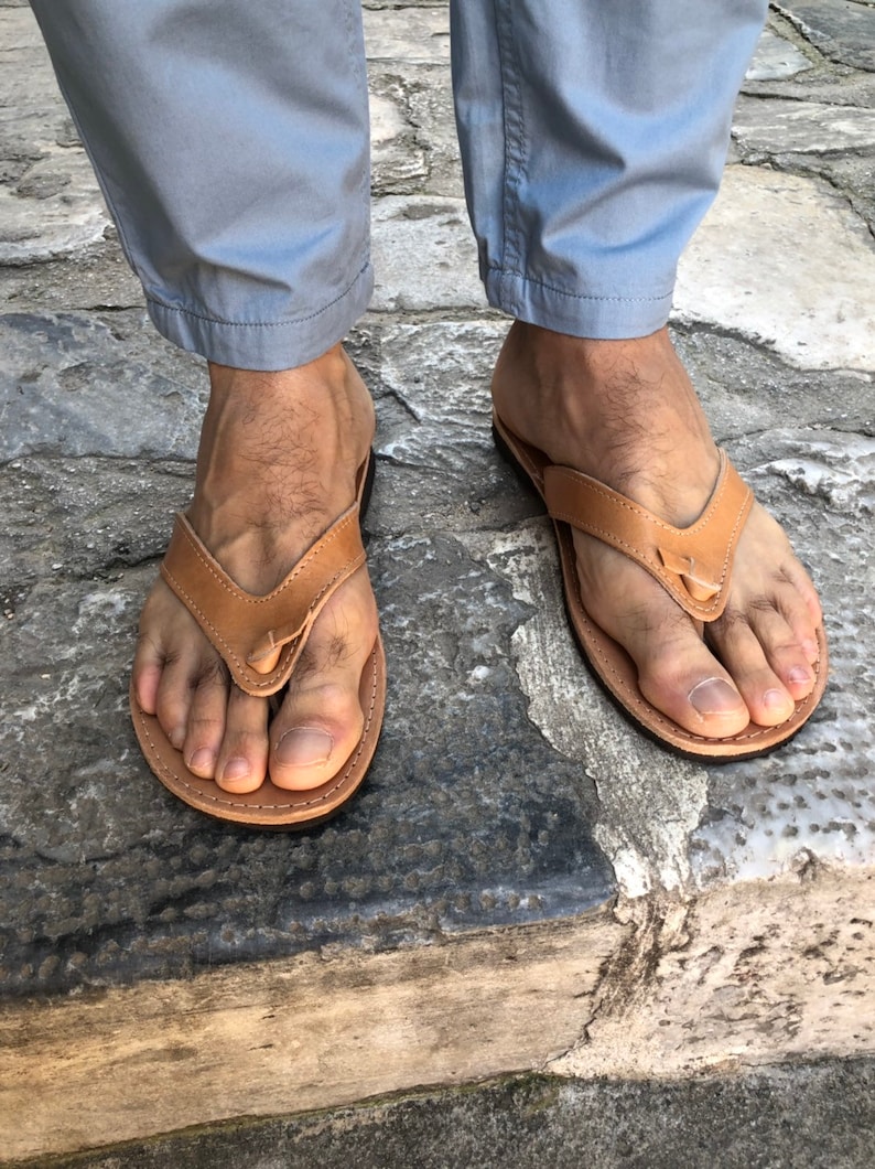 Pode incluir: Um par de chinelos de couro castanho claro. As sand&aacute;lias t&ecirc;m um design de tanga com uma tira entre os dedos. As solas s&atilde;o castanhas mais escuras. As sand&aacute;lias s&atilde;o usadas com cal&ccedil;as cinza claro.