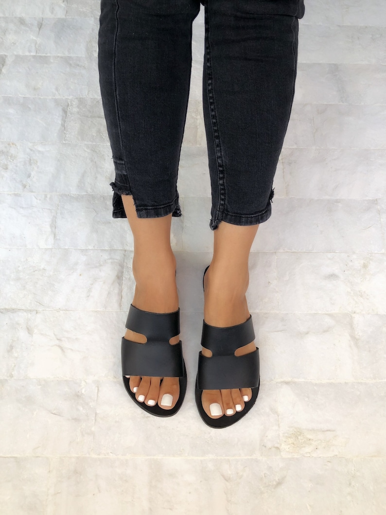 Puede incluir: Sandalias de cuero negro con un dise&ntilde;o minimalista. Las sandalias presentan dos correas anchas en la parte superior del pie y una suela plana. La imagen muestra las sandalias con unos vaqueros negros.