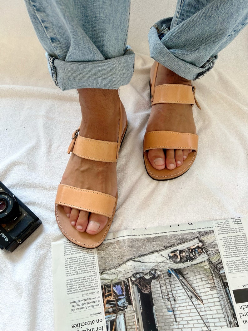 May include: A pair of tan leather sandals with two straps and a buckle closure. The sandals are worn by a person with bare feet and are resting on a white surface.