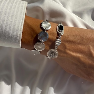 May include: Silver dangle earrings with geometric shapes. One earring features a black rectangular stone. The other earring has three circular discs. The earrings are displayed against a white background.