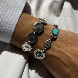 May include: Two silver bracelets. One has hammered, circular links. The other features a turquoise-coloured rectangular stone and geometric shapes. The bracelets are worn on a wrist, against a white shirt.
