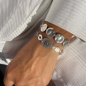 May include: Two silver bracelets with unique designs. One bracelet features a series of hammered, circular charms. The other bracelet has a mix of circular and patterned elements. The bracelets are worn on a wrist, against a white shirt.