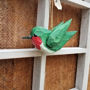 May include: A green and red wooden hummingbird ornament hanging from a white wooden frame. The ornament has a small tag that reads "Songbird Creations - Handcrafted in Texas".