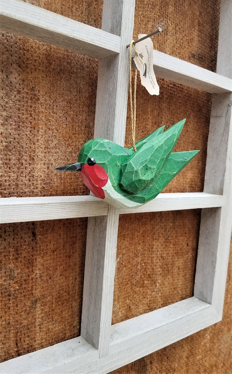May include: A green and red wooden hummingbird ornament hanging from a white wooden frame. The ornament has a small tag that reads "Songbird Creations - Handcrafted in Texas".