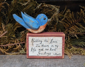 Puede incluir: Una figurita de pájaro azul de madera posada sobre un pequeño bloque de madera con el texto "Holding the love I've known in my life and no hard feelings - Avett" pintado en él.
