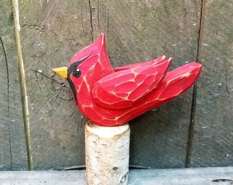 May include: A hand-carved wooden cardinal bird figurine painted red with black eyes and a yellow beak. The bird is perched on a small white birch log.