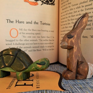 May include: A wooden toy hare and a wooden toy turtle, both painted in realistic colors. The hare is brown with a white patch on its side. The turtle is green with a brown shell. The toys are sitting on a book with the text "The Hare and the Tortoise" visible.