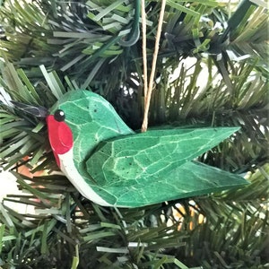 May include: A green wooden hummingbird ornament with a red beak and black eyes. The ornament is hanging from a brown string on a green artificial Christmas tree.