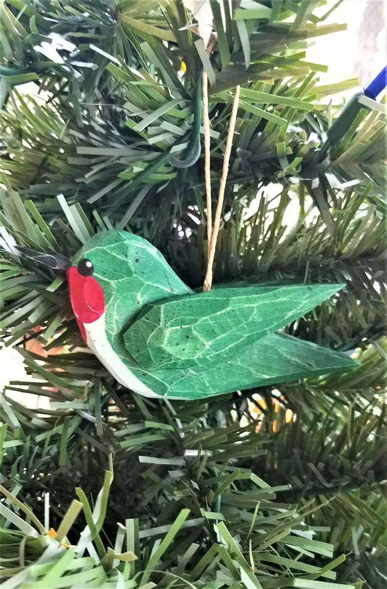 May include: A green wooden hummingbird ornament with a red beak and black eyes. The ornament is hanging from a brown string on a green artificial Christmas tree.