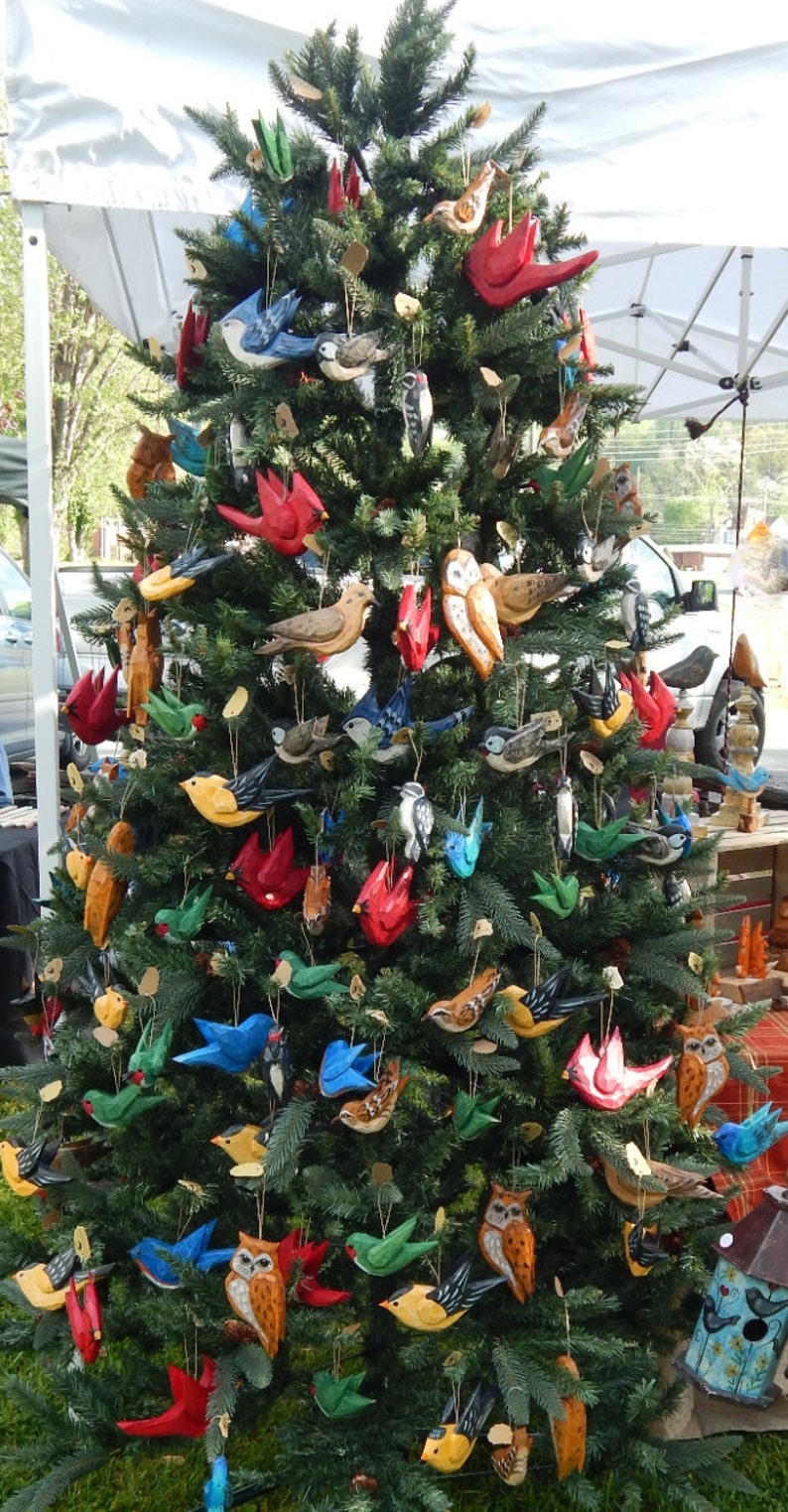 May include: A Christmas tree decorated with hand-painted wooden ornaments. The ornaments feature a variety of birds, owls, and other woodland creatures. The ornaments are painted in bright colors, including red, blue, green, yellow, and brown.