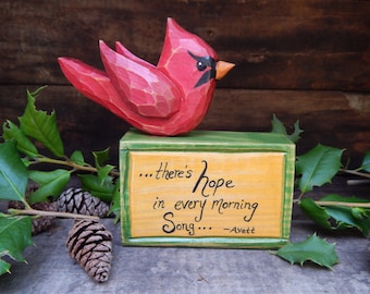 May include: A wooden carved cardinal bird perched on a small wooden box with a green top. The box has a yellow background with the text "...there's hope in every morning song... -Avett" written in black.