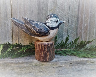 May include: A hand-carved wooden bird figurine with black and white painted details, perched on a small wooden stump. The bird is facing the right side of the image and has a realistic appearance.