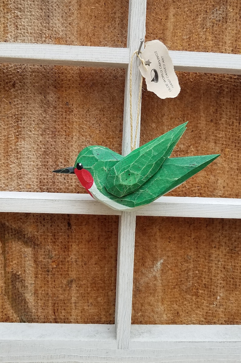 May include: A green wooden hummingbird ornament with red accents. The ornament is hanging from a white wooden frame with a tag that reads "Olde World Carvings Hand Carved Art".