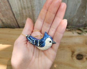 Op de afbeelding: Een ketting met een hanger in de vorm van een blauwe gaaien op een gouden ketting. De vogel is geschilderd met witte en zwarte details. De hanger is gemaakt van hout en is een realistische afbeelding van een blauwe gaaien.