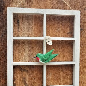 May include: A white wooden window frame with four panes, hanging on a brown burlap background. A green and red wooden hummingbird ornament is hanging from the top pane.