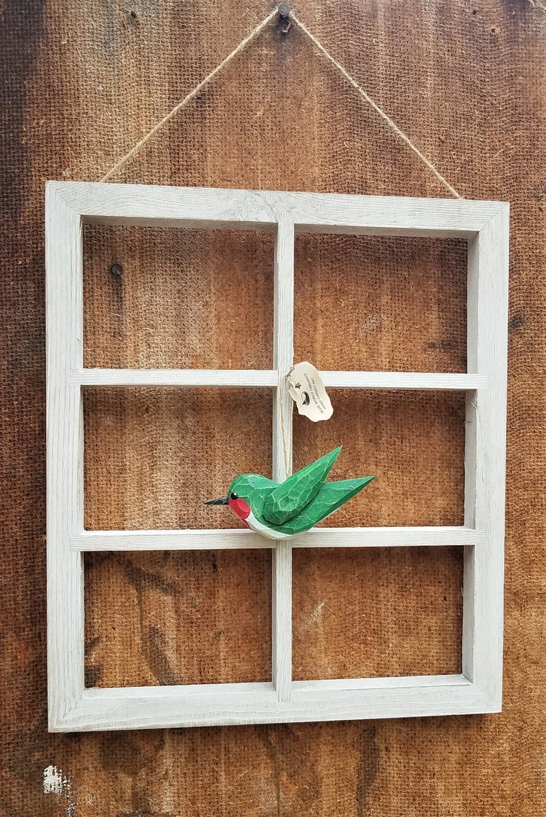 May include: A white wooden window frame with four panes, hanging on a brown burlap background. A green and red wooden hummingbird ornament is hanging from the top pane.