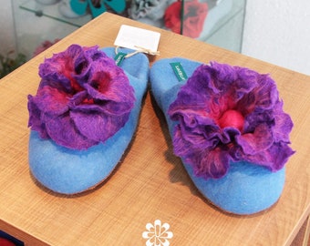 May include: A pair of blue felt slippers with purple and pink felt flowers on top. The slippers are on a wooden surface with the text "artslippers" printed on it.