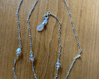 Könnte beinhalten: Eine silberne Kette mit klaren Kristallperlen, die zum Halten von Brillen gedacht ist. Die Kette hat an den Enden zwei Silikon-Schlaufen zum Befestigen an Brillen.