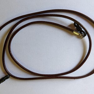 Può includere: Un cordino per occhiali in pelle marrone con anelli in gomma nera e dettagli in metallo argentato. Il cordino è ad anello su una superficie bianca. Il cordino è progettato per tenere gli occhiali al sicuro e accessibili.