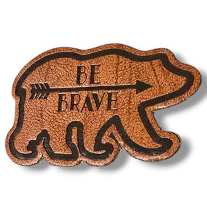 Könnte beinhalten: Ein brauner Leder-Bären-Patch mit schwarzem Umriss. Der Bär hat einen Pfeil, der nach rechts zeigt, mit den Worten "BE BRAVE" darunter.