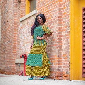 May include: A woman wearing a green and yellow patterned dress with a ruffled neckline and sleeves. She is standing in front of a brick wall with a yellow door.