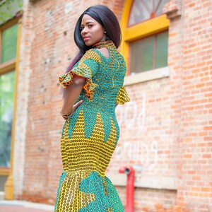 May include: A woman wearing a yellow and blue patterned maxi dress with a ruffled hem. The dress has a high neck and short sleeves. The woman is standing in front of a brick wall.
