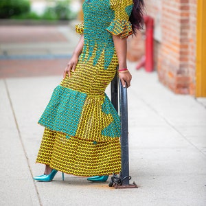 May include: A woman wearing a yellow and blue patterned dress with a ruffled skirt. She is standing next to a black metal pole.