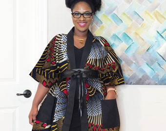 May include: A colorful patterned kimono-style jacket with black trim and a black belt. The jacket features red, yellow, and blue designs on a black background. The jacket has two black pockets and is worn over a black dress.