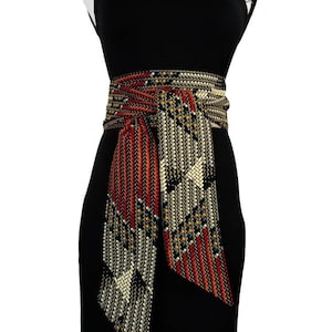 Könnte beinhalten: Ein schwarzes Kleid mit einem dekorativen gewebten Gürtel. Der Gürtel hat ein geometrisches Muster in Rot-, Gold- und Cremefarben. Das Kleid hat ein ärmelloses Design mit dekorativen Schulterträgern.