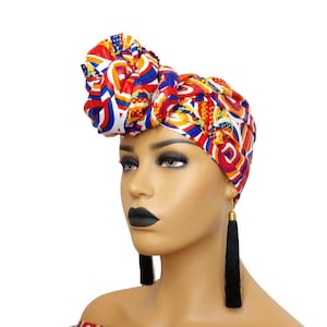 May include: A woman wearing a colourful head wrap with a geometric pattern. The wrap is tied in a knot on top of her head. She is also wearing long black tassel earrings.