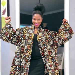 Op de afbeelding: Een lange, patroon kimono-stijl jas over een zwarte jurk. De jas heeft een levendige mix van geometrische en dierenprints in zwart, geel, oranje en wit. De mouwen zijn wijd en vloeiend, wat een gedurfd silhouet creëert. De stijl is een mix van moderne en etnische invloeden.