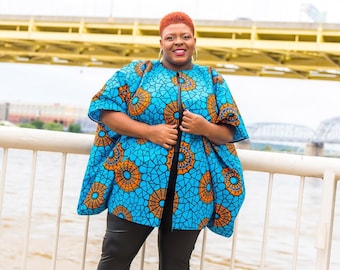 May include: A woman wearing a blue and orange patterned jacket with a black zipper, black leggings, and black heeled boots. She is standing in front of a white railing with a river and a bridge in the background.