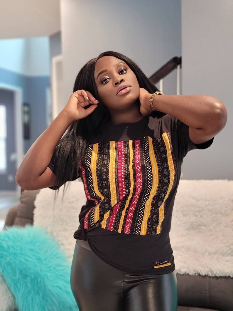 May include: A black t-shirt with a colorful geometric pattern. The pattern features stripes of yellow, red, and black with intricate designs.