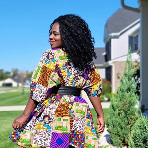 May include: A vibrant patchwork dress with a black belt. The dress showcases a mix of patterns and colours, such as yellow, purple, and brown. It is worn with black leggings. The background includes a house, green grass, and a clear blue sky.