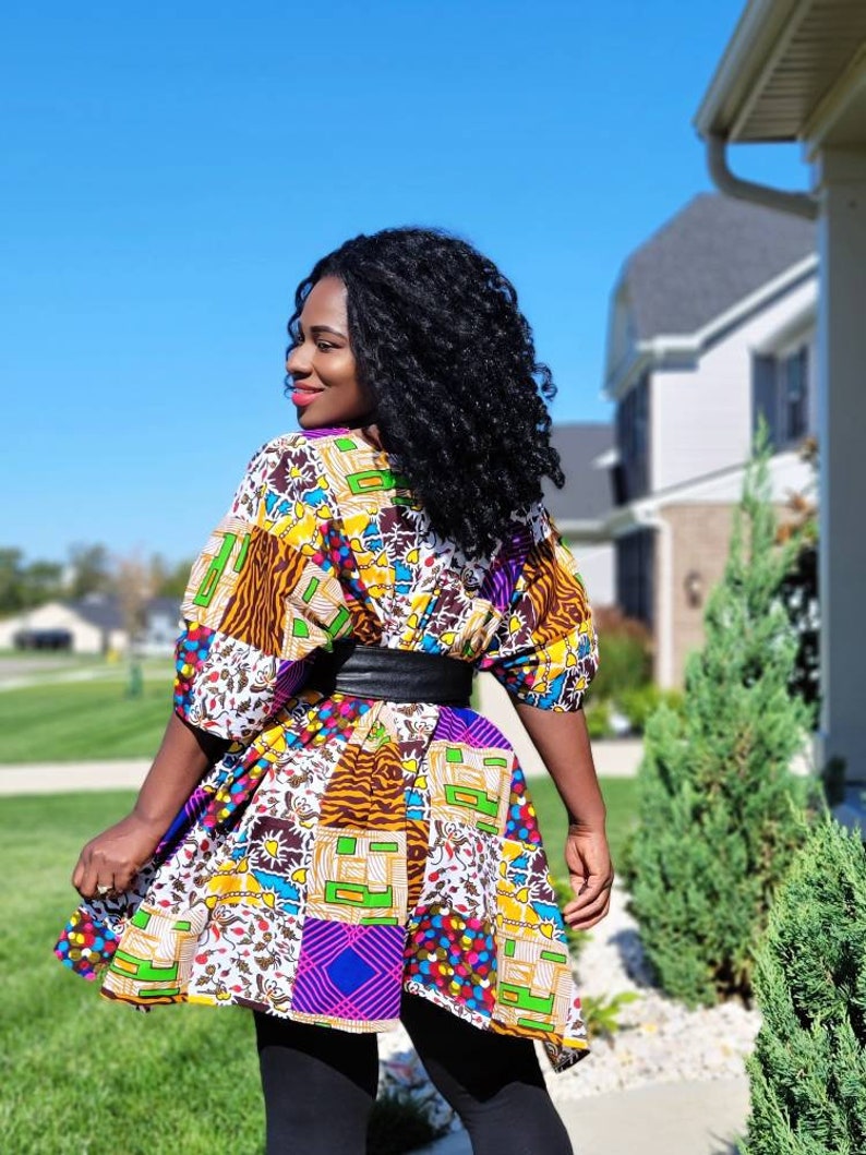 May include: A vibrant patchwork dress with a black belt. The dress showcases a mix of patterns and colours, such as yellow, purple, and brown. It is worn with black leggings. The background includes a house, green grass, and a clear blue sky.