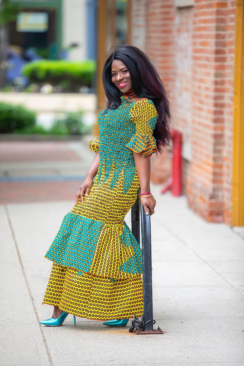 May include: A woman wearing a yellow and blue patterned dress with a ruffled skirt. She is standing next to a black metal pole.
