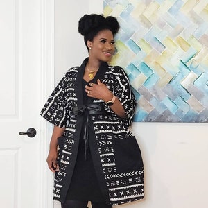 May include: A black and white patterned kimono with a black dress and black leggings. The kimono has a geometric pattern with white accents. The person is wearing a gold necklace with an Africa-shaped pendant.