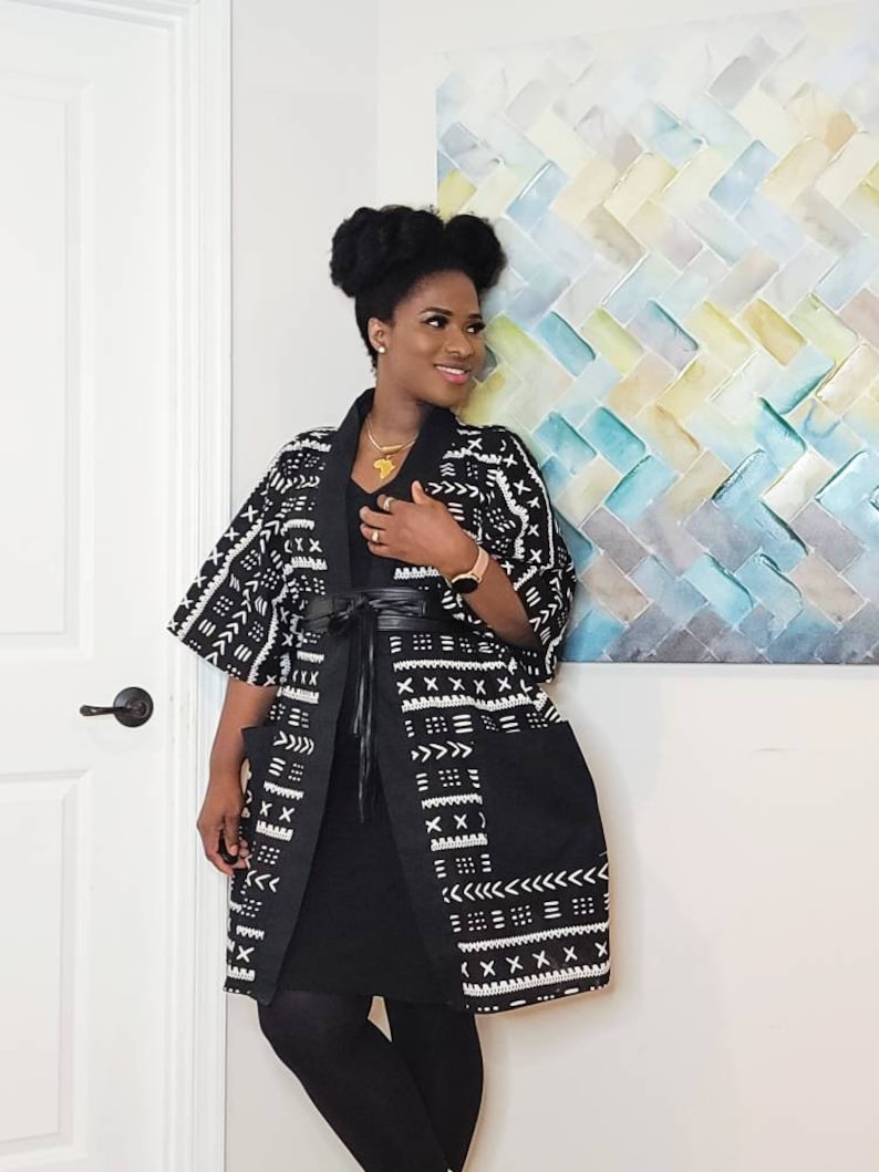 May include: A black and white patterned kimono with a black dress and black leggings. The kimono has a geometric pattern with white accents. The person is wearing a gold necklace with an Africa-shaped pendant.