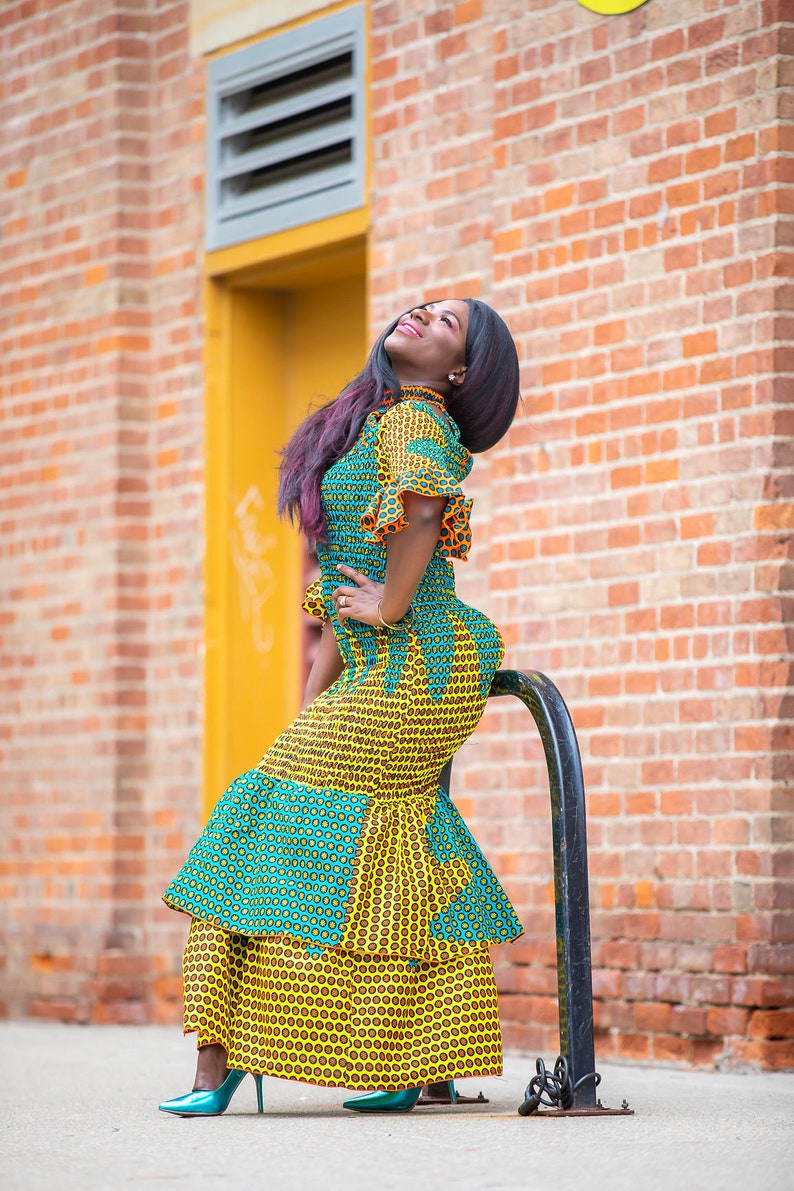 May include: A woman wearing a yellow and green patterned dress with a ruffled skirt. She is standing in front of a brick wall and leaning against a black metal pole.