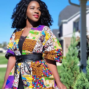 May include: A vibrant patchwork kimono jacket with a black belt. The jacket showcases diverse patterns, including floral, geometric, and animal prints, in purple, yellow, blue, and brown. The model is wearing a black top underneath.
