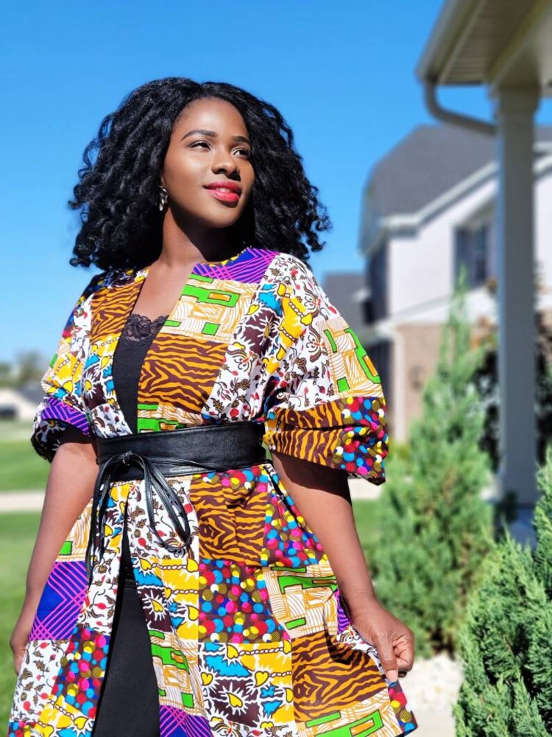 May include: A vibrant patchwork kimono jacket with a black belt. The jacket showcases diverse patterns, including floral, geometric, and animal prints, in purple, yellow, blue, and brown. The model is wearing a black top underneath.