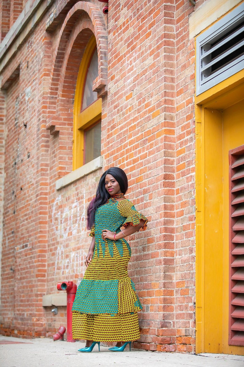 May include: A woman wearing a green and yellow patterned dress with a ruffled neckline and sleeves. She is standing in front of a brick wall with a yellow door.