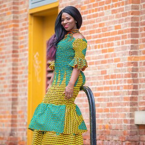 May include: A woman wearing a yellow and green patterned dress with a ruffled skirt. She is standing in front of a brick wall and holding onto a black metal pole.