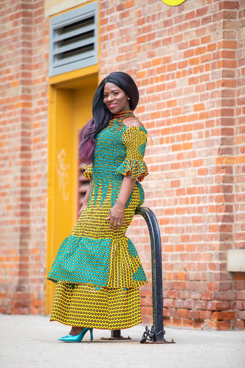 May include: A woman wearing a yellow and green patterned dress with a ruffled skirt. She is standing in front of a brick wall and holding onto a black metal pole.