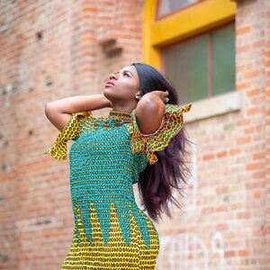 May include: A woman wearing a yellow and blue patterned dress with ruffled sleeves and a high neckline. She is standing in front of a brick wall with a window behind her.