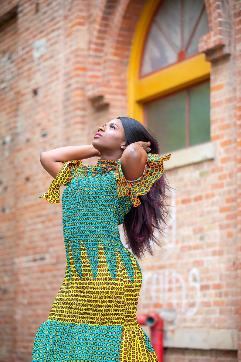May include: A woman wearing a yellow and blue patterned dress with ruffled sleeves and a high neckline. She is standing in front of a brick wall with a window behind her.