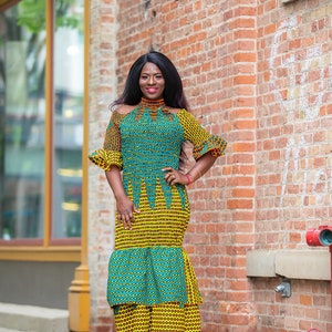 May include: A woman wearing a green and yellow patterned dress with a high neckline and short sleeves. The dress has a tiered skirt and the woman is standing in front of a brick wall.