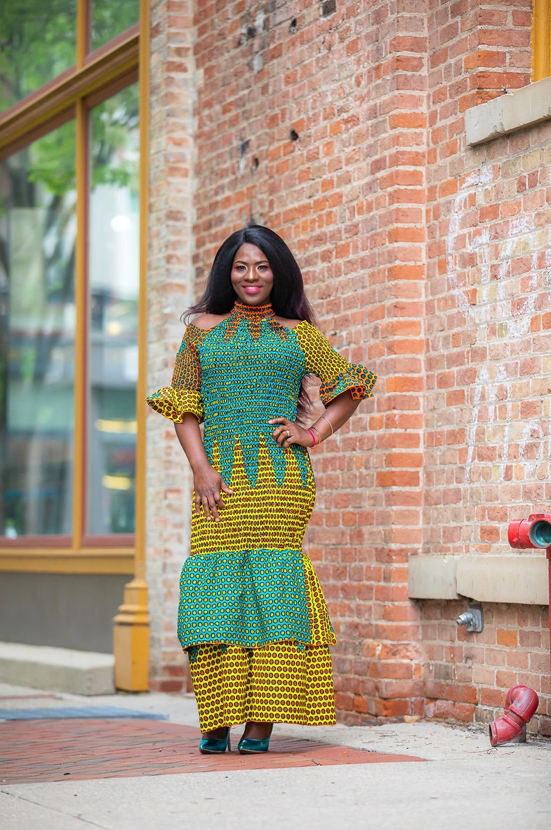 May include: A woman wearing a green and yellow patterned dress with a high neckline and short sleeves. The dress has a tiered skirt and the woman is standing in front of a brick wall.