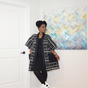 May include: A woman wearing a black dress with a white and black patterned kimono. She is wearing white ankle boots and a gold necklace.